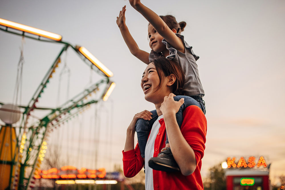 Mother and child at a carnival at dusk, smiling as the child waves from her shoulders.